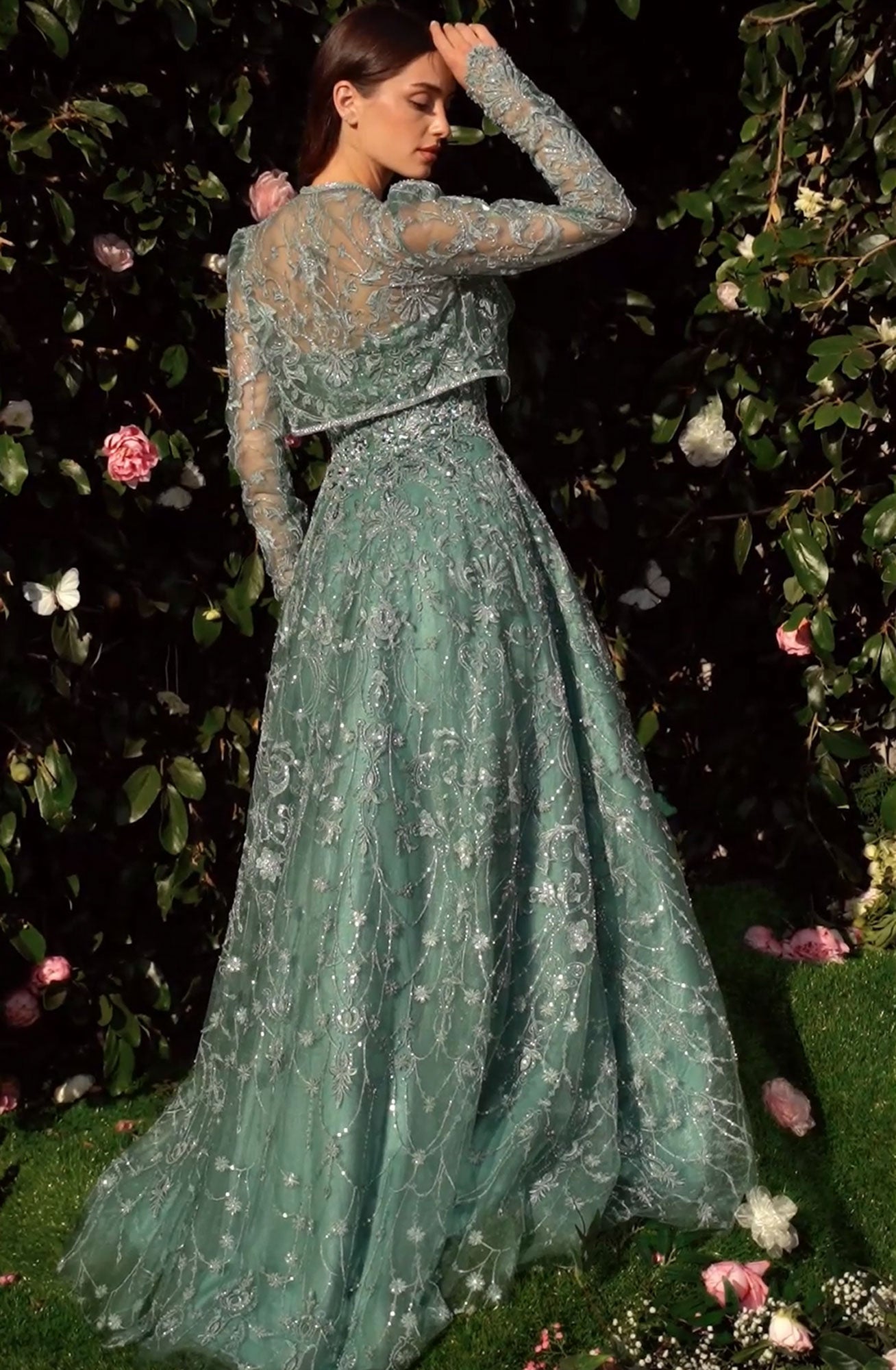 A woman stands in a garden wearing the Andrea & Leo MA104 Long Sleeve Long Formal Dress with embroidered green lace, facing away and touching her head as pink and white flowers bloom in the background foliage.